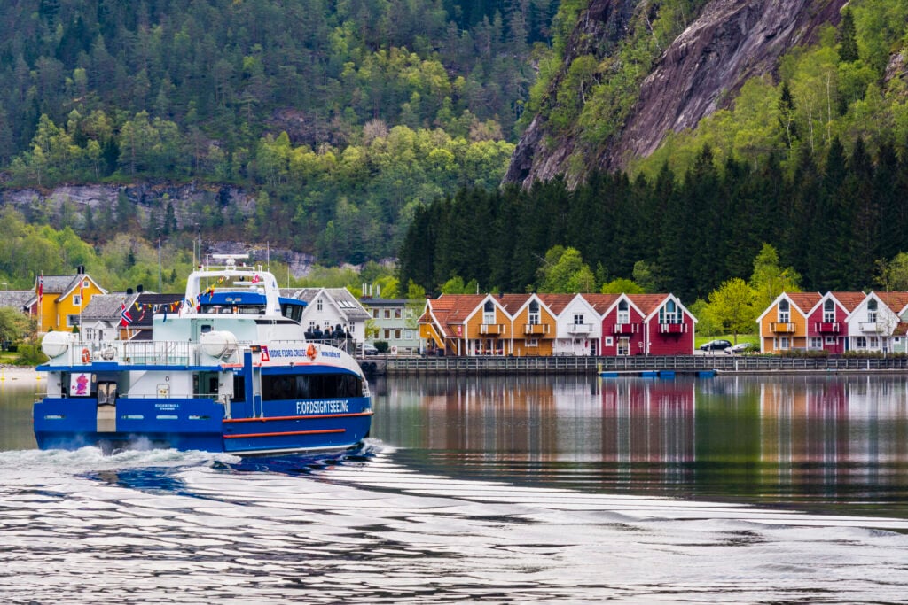 A small modern fjord cruise catamaran gliding past colorful wooden houses along the shoreline, with green hills and forests in the background near Bergen, Norway.