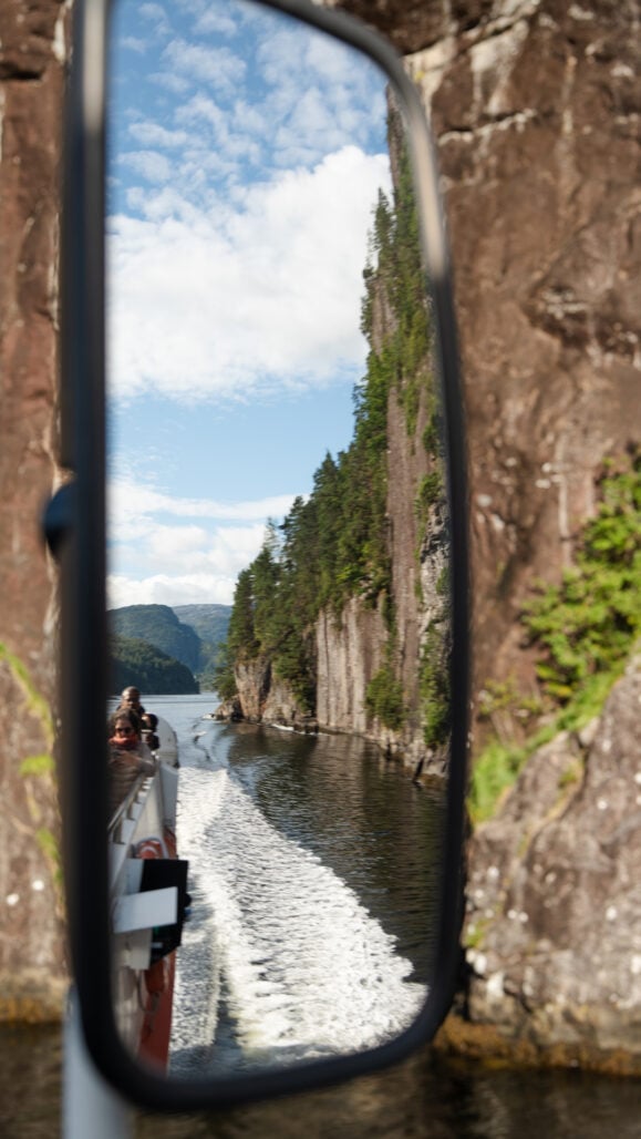 Guests reflected in a boat mirror enjoying the view of Fugleberget and the surrounding fjord during a Mostraumen cruise.