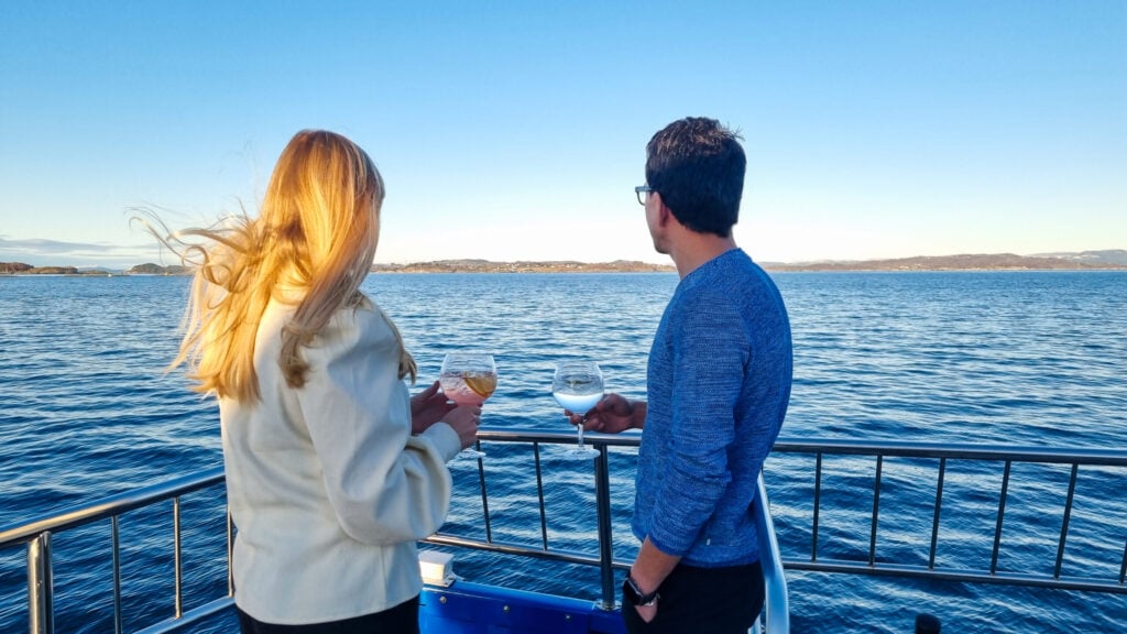 Par nyter ginsmaking på dekk under fjordcruise fra Stavanger, med utsikt over fjorden og solskinn i bakgrunnen.