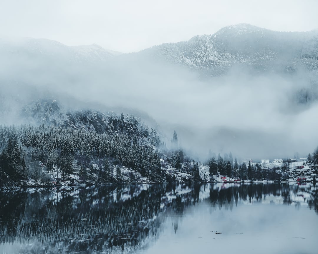 Misty winter landscape with snow-covered forest and mountains reflected in calm waters at Mostraumen near Bergen