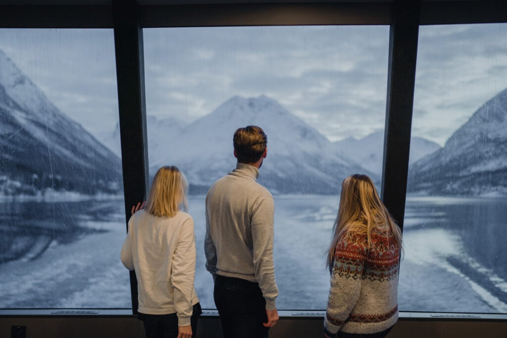 3 reisende nyter utsikten fra de store panomarma vinduene på et fjordcruise fra Tromsø. Med Lyngenalpene i bakgrunnen