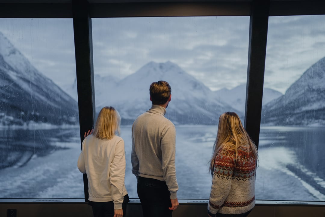 3 reisende nyter utsikten fra de store panomarma vinduene på et fjordcruise fra Tromsø. Med Lyngenalpene i bakgrunnen