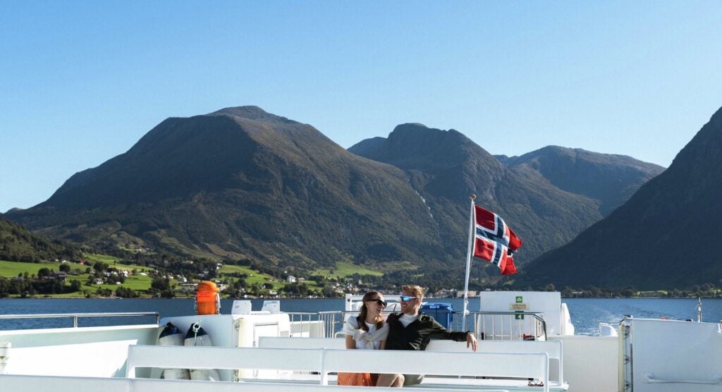 Par som nyter fjordcruise i fra Bergen til Rosendal med norsk flagg og fjell i bakgrunnen