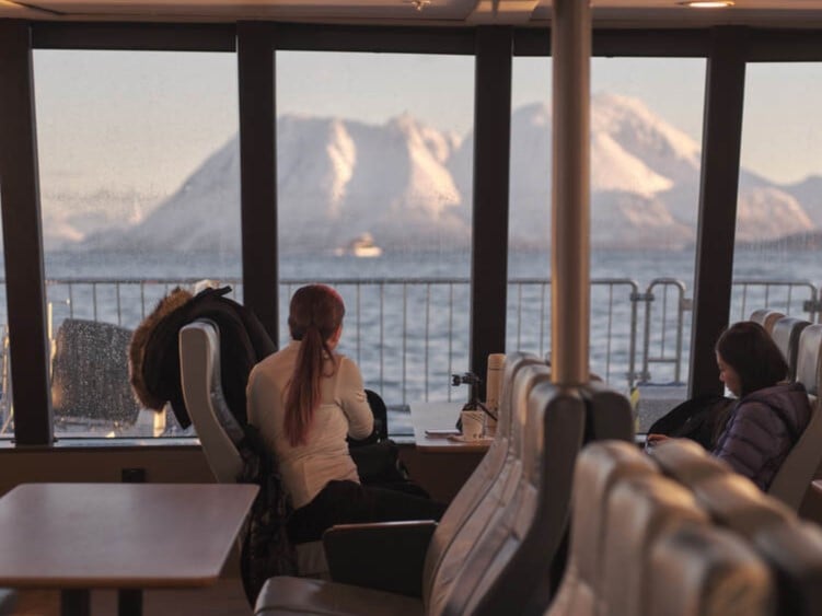 Vinterlandskap sett fra store panoramiske vinduer i salongen på fjordcruise i Tromsø, med snødekte fjell i bakgrunnen.