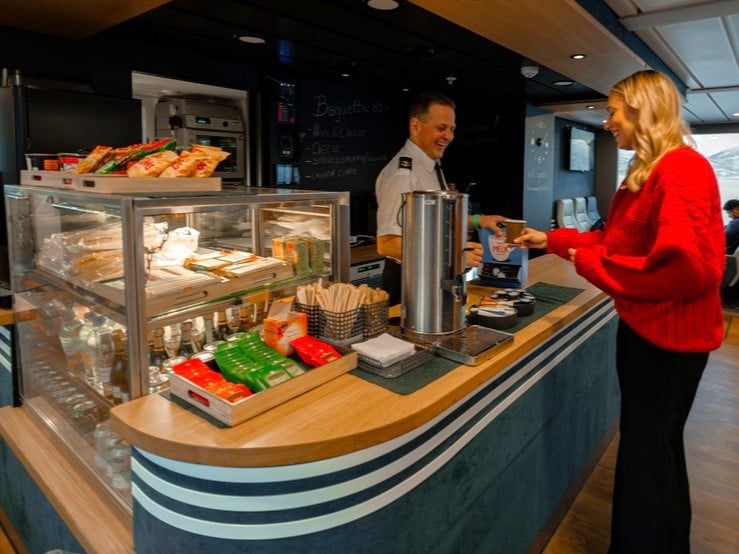 Ein lächelnder Fahrgast in einem roten Strickpullover kauft Snacks und warme Getränke von der Besatzung in dem geräumigen Kiosk an Bord einer Nordlicht-Kreuzfahrt in Tromsø. Ein angenehmes und komfortables Erlebnis.