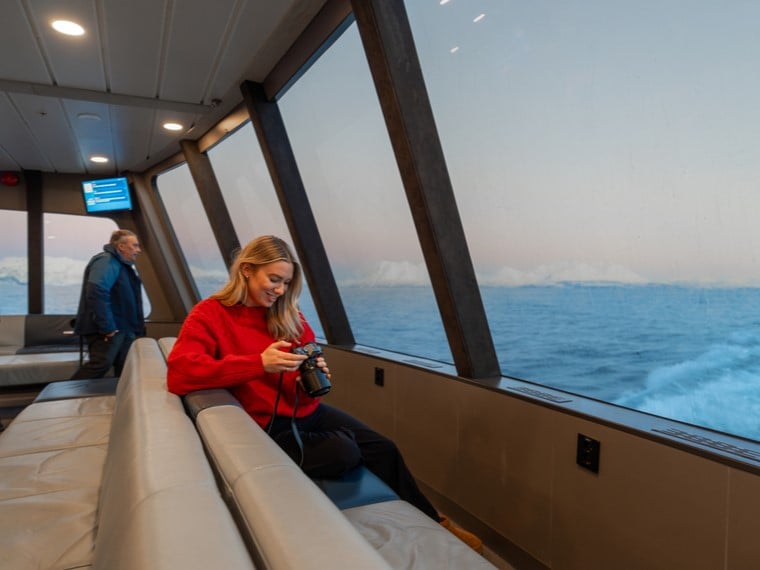 En kvinne sitter komfortabelt i kabinen på fjordcruiset i Tromsø, omgitt av store panoramavinduer. Hun ser gjennom bildene fra turen mens majestetiske fjorder og snødekte fjell glir forbi.
