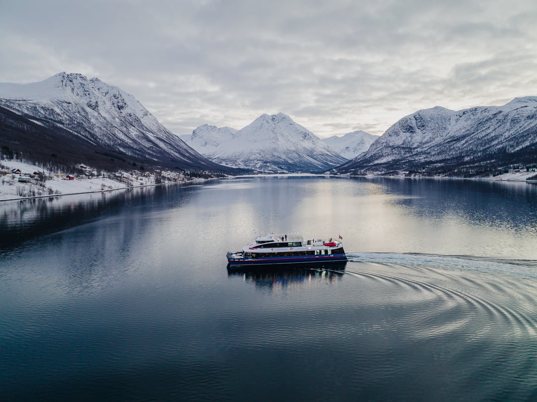 Drone bilde av Rygerdronningen på fjord cruise i Tromsø. Med snøkledde fjell og nord norsk natur i bakgrunnen.