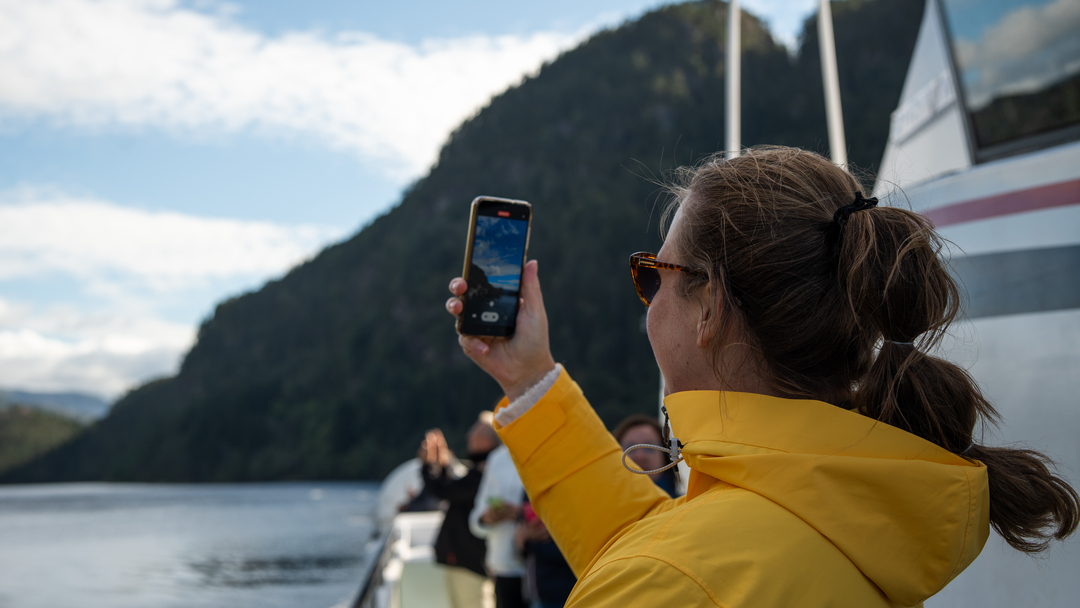Kvinne i gul jakke som koser på båttur med Rødne Fjord Cruise fra Bergen og står ute blant flere folk og filmer det vakre fjordlandskapet.