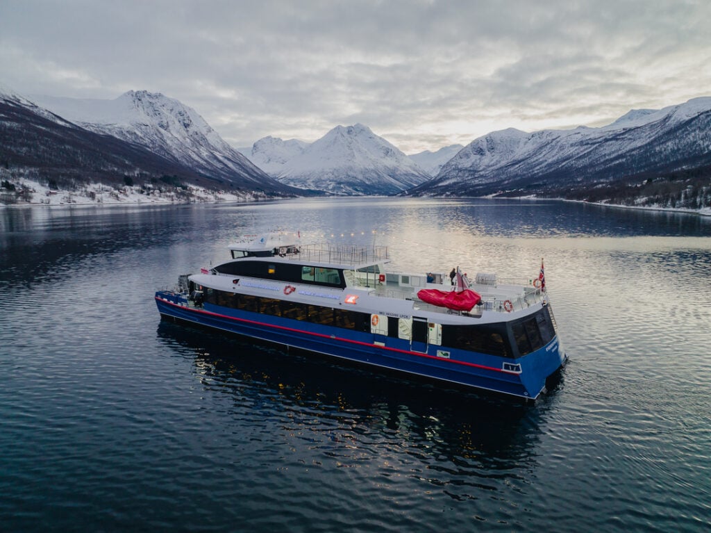 Aerial view of Rødne’s boat Rygerdronningen cruising through an Arctic fjord near Tromsø, surrounded by snow-covered mountains and calm winter waters.