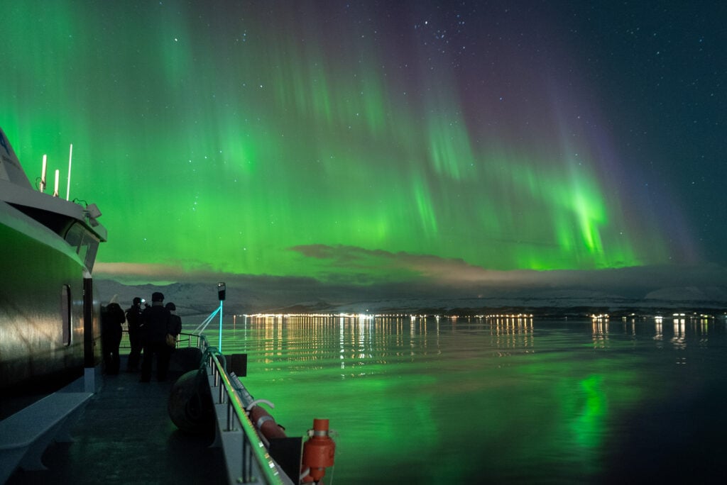 Guests on the boat deck taking photos as Tromsø is illuminated by green Northern Lights, reflecting on the Arctic waters.