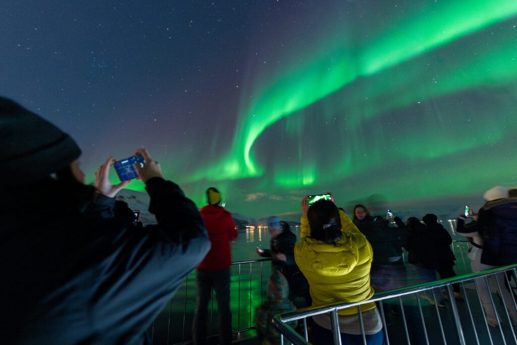 Guests on the deck of a Rødne boat taking photos of the green Aurora Borealis dancing above the Arctic fjord near Tromsø.
