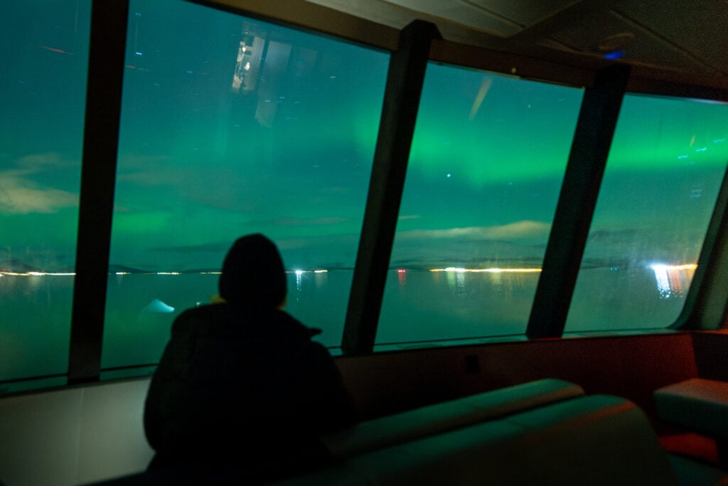 Guest watching the Northern Lights through large panoramic windows on board, with Tromsø’s city lights glowing across Tromssundet.
