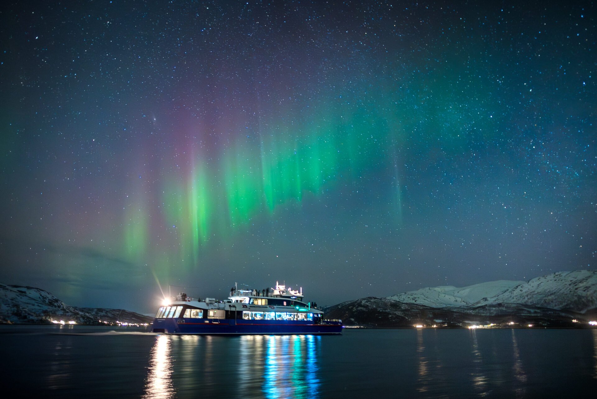 Rødne’s boat Rygerdronningen sailing under the Northern Lights outside Tromsø, with the city lights glowing in the background.