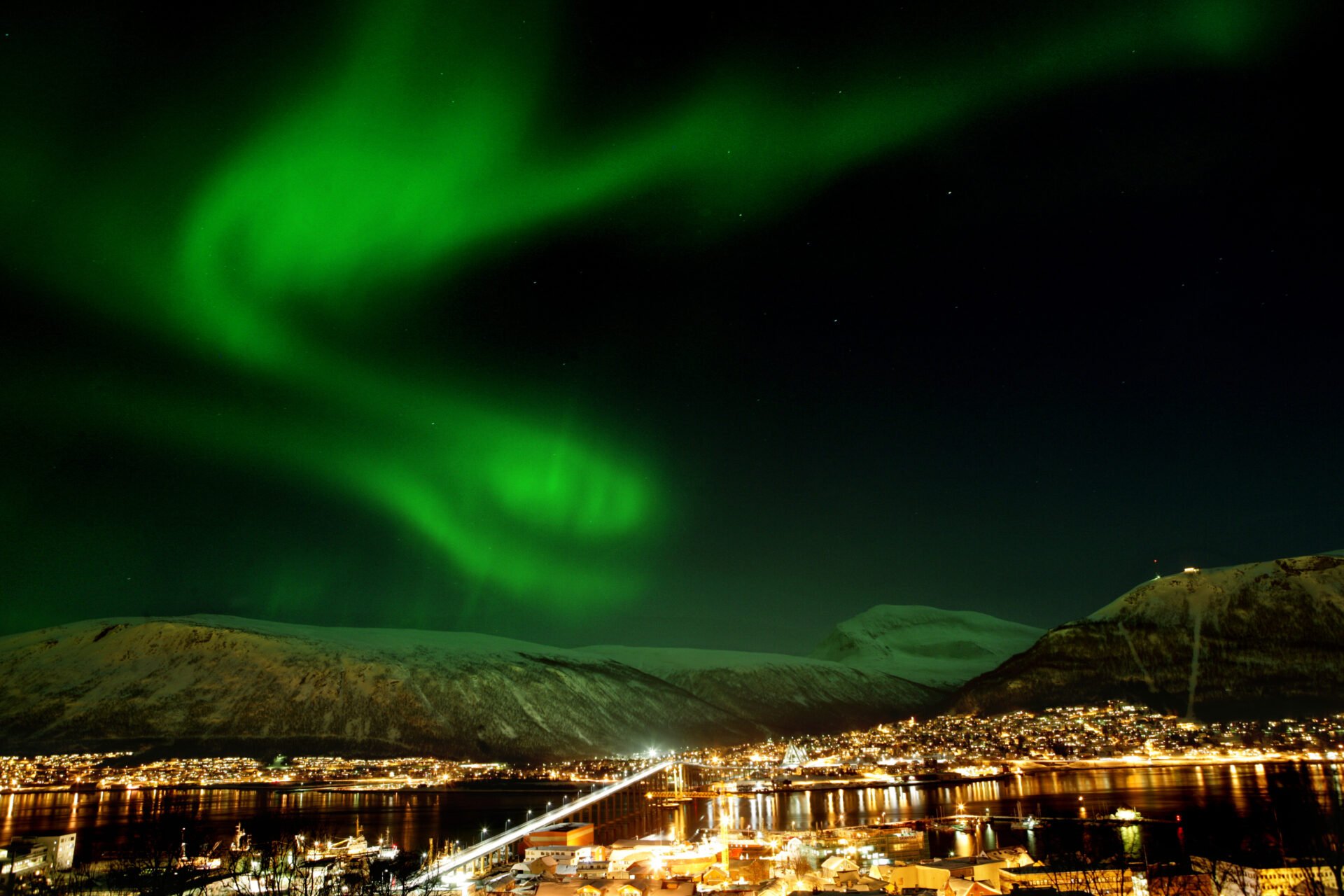 Northern Lights dancing over Tromsø city at night, with glowing city lights and Arctic fjords surrounding the island.