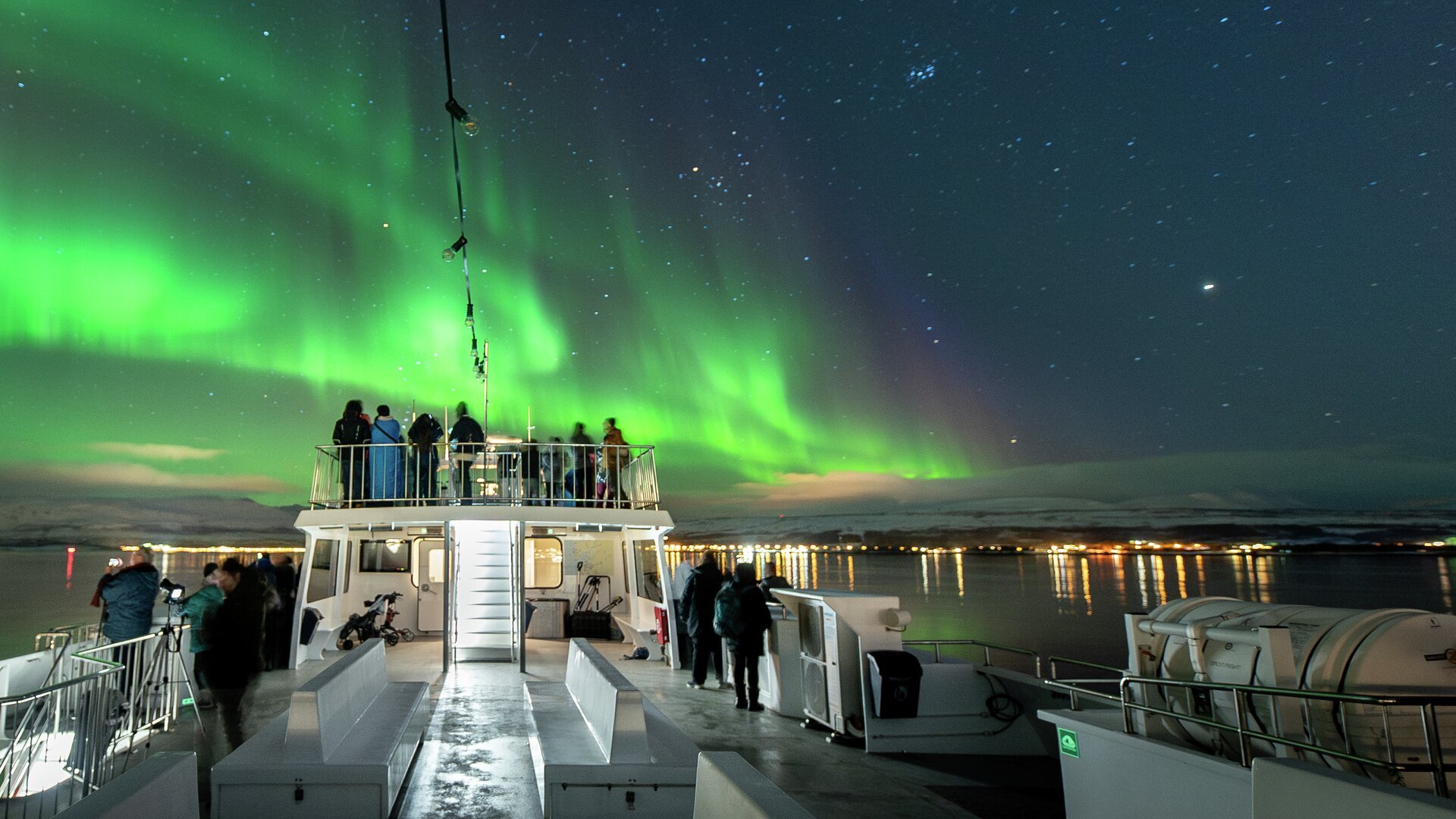 Guests in winter clothing taking photos of the Northern Lights from the deck, with Tromsø glowing in the distance under a starry sky.