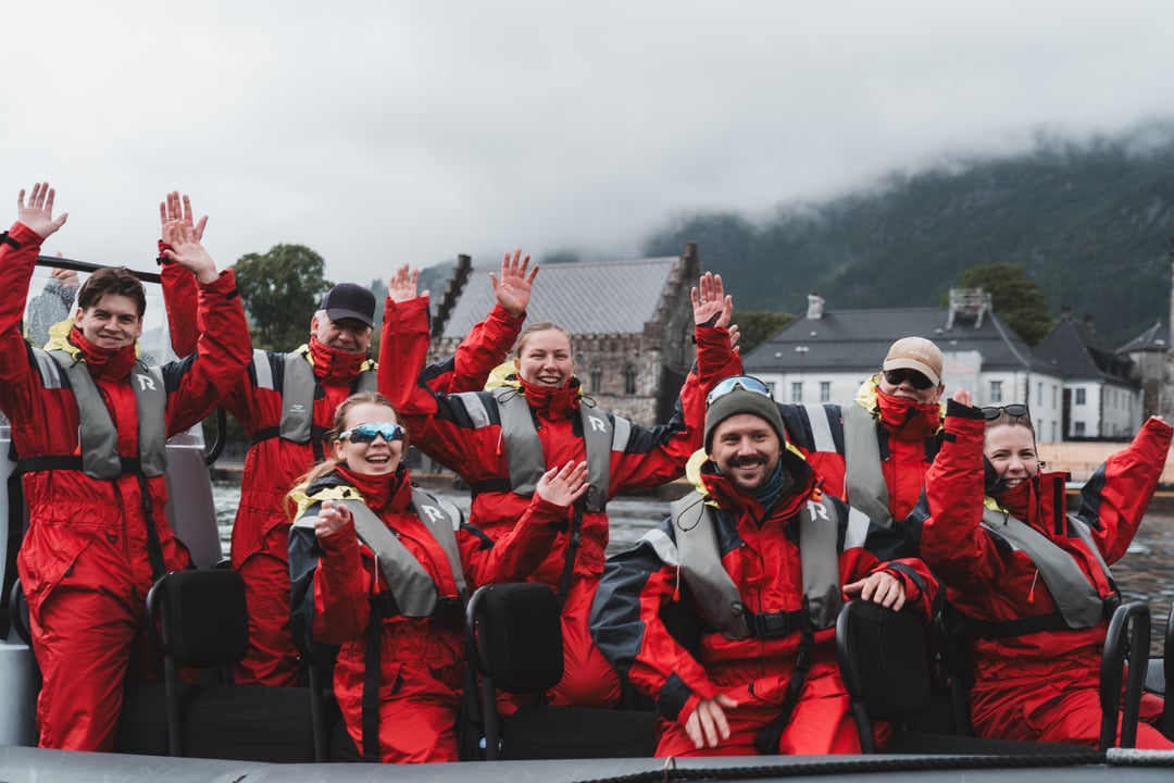 Smiling RIB guests in red protective suits waving, with the historic Bergenhus fortress behing.