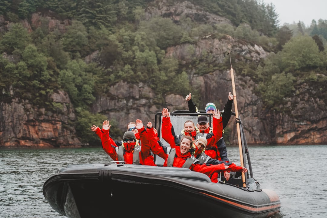 RIB passengers in red suits cheering with the steep green cliffs behind, west of Bergen.
