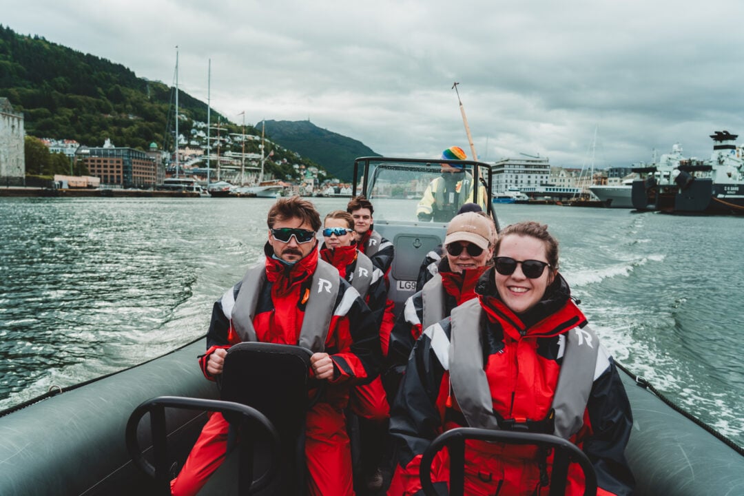 Smiling RIB guiest in red Rødne flotation suits cruising Bergen Harbour with the city and mountains behind.