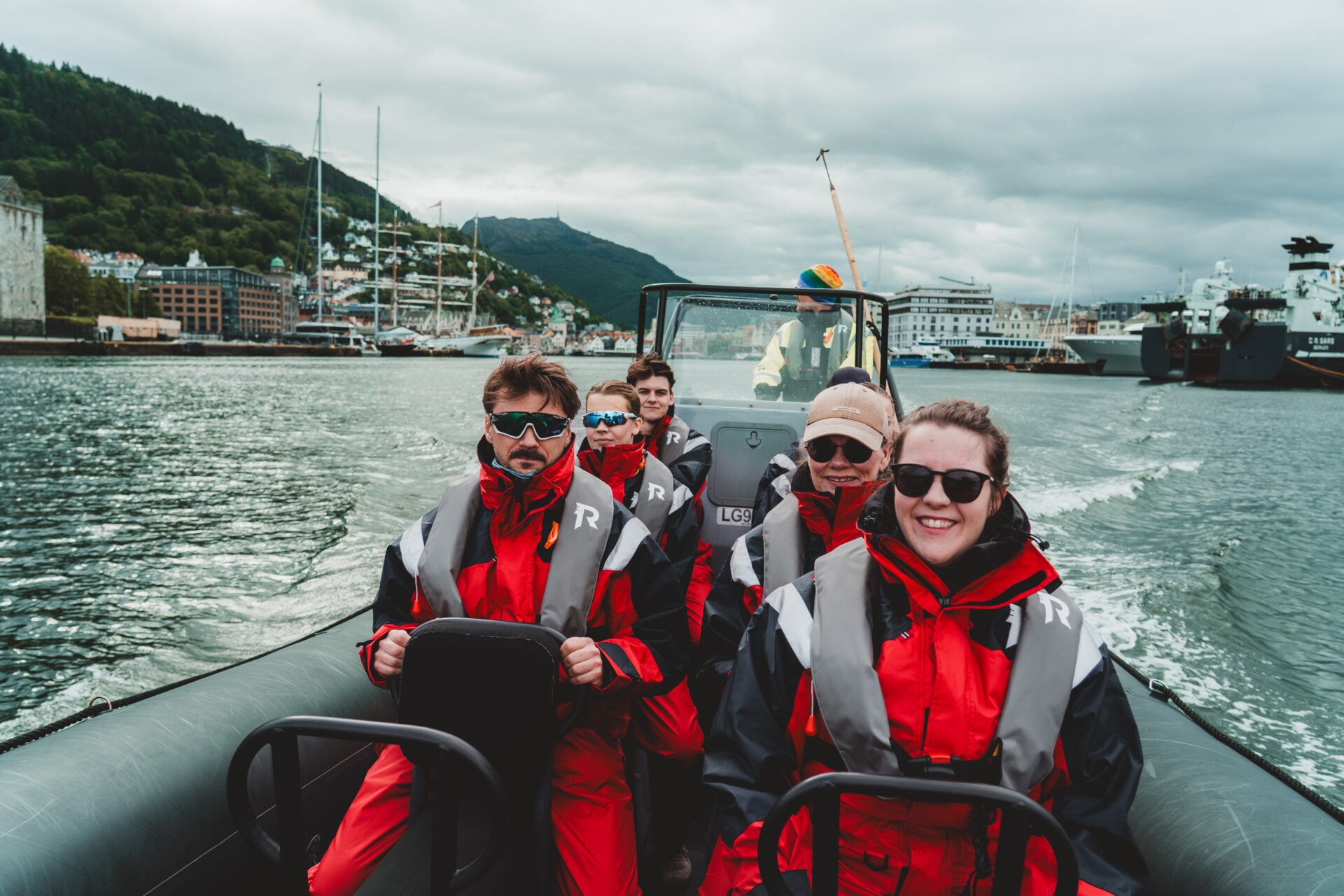 Smiling RIB guiest in red Rødne flotation suits cruising Bergen Harbour with the city and mountains behind.