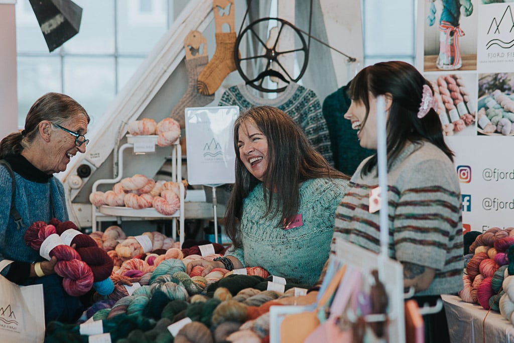 Kvinner som selger fargerikt garn på Bergen Strikkefestival som ler og koser seg i dialog med festivalgjest og potensiell kjøper. Markedet til Bergen Strikkefestival.