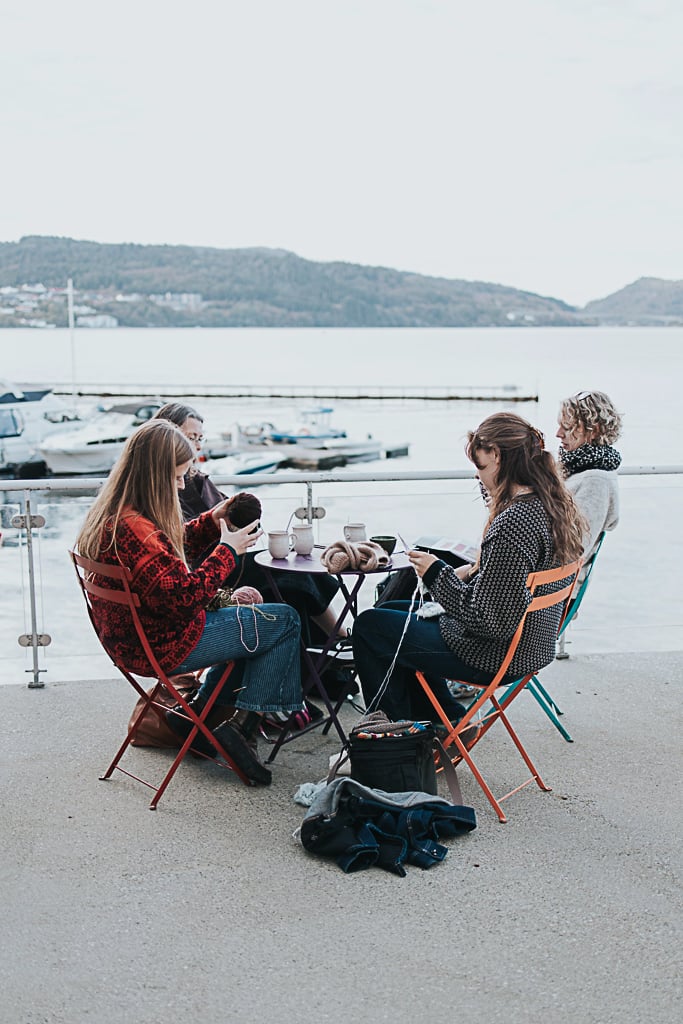 Fire kvinner i strikkegensere sitter rundt et rundbord på caféstoler og strikker og prater på kaien ved fjorden, utenfor Trikotasjemuseet i Salhus.